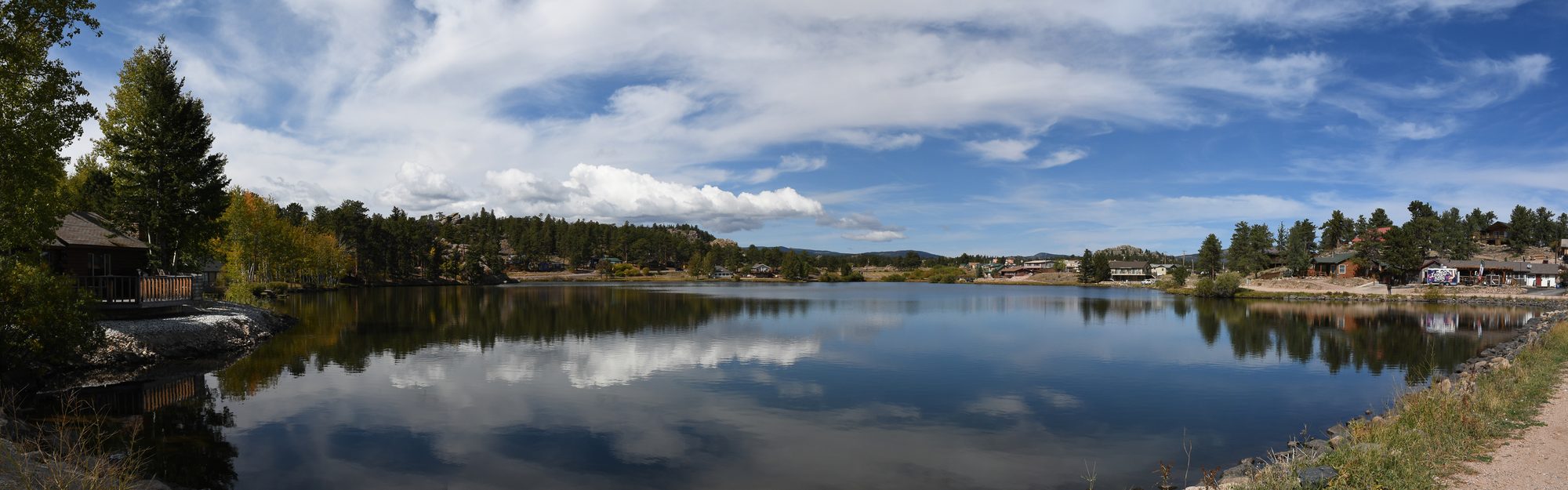 A calm lake reflecting clouds, ringed by pines and small cabins along the shore
