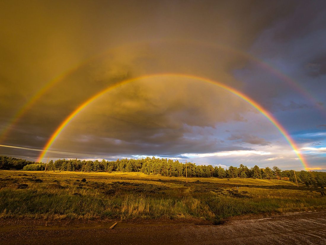 A full double rainbow arches over a grassy field and tree line after rain