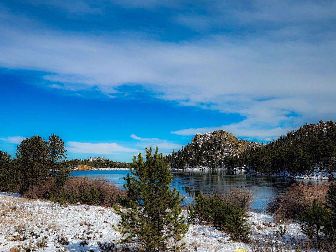 A partly frozen lake with snowy banks, scattered pines and a rocky outcrop under blue sky