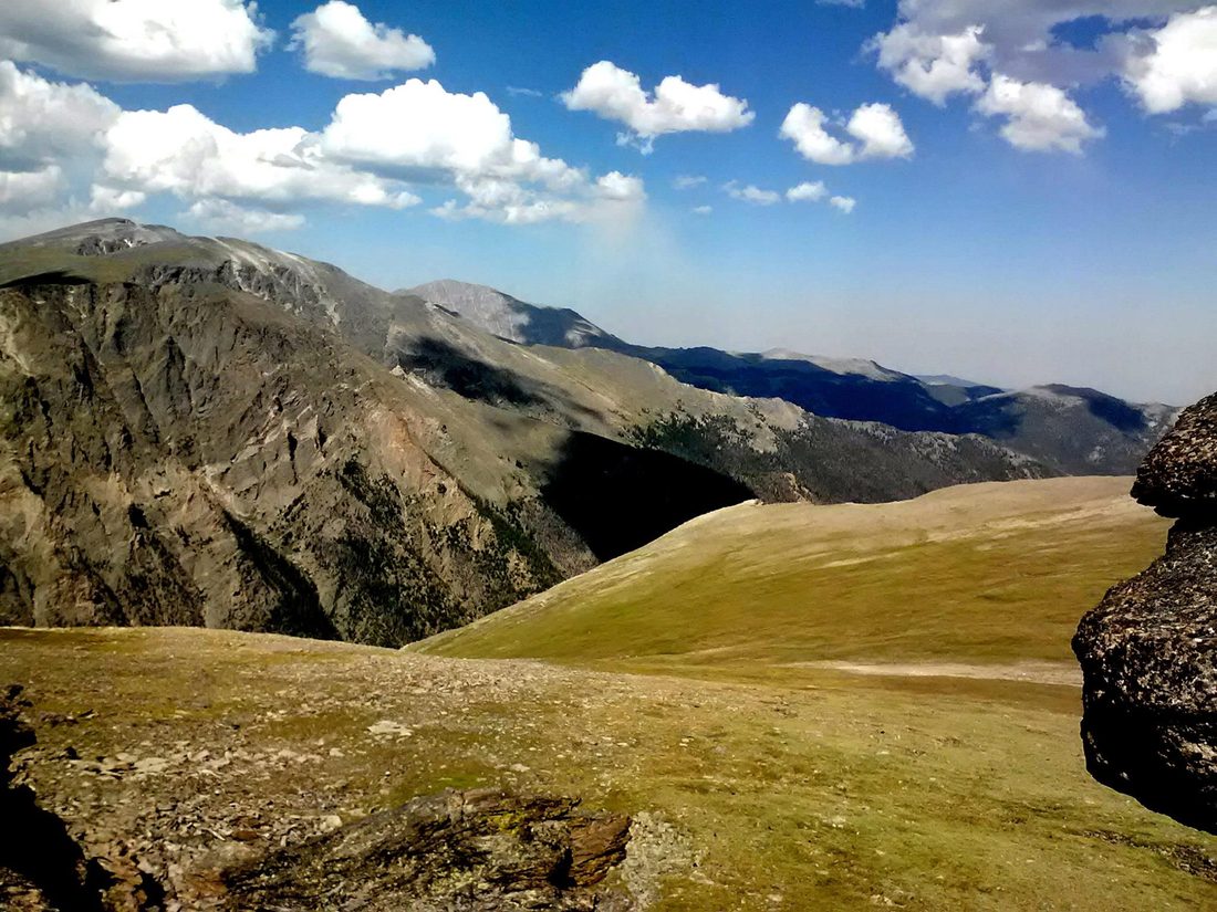 Rocky mountain ridges and a grassy high-country basin under a bright cloud-dotted sky