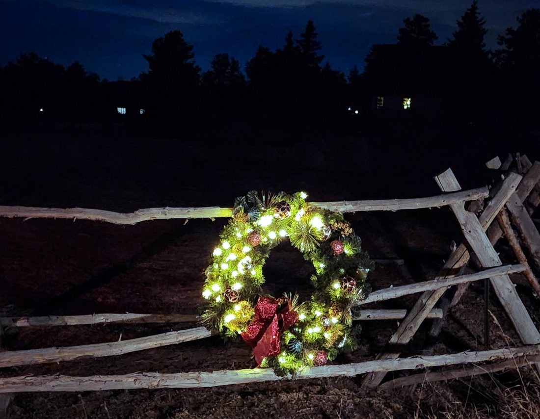 A glowing lit holiday wreath with a red bow leans on a rustic wooden rail fence at night