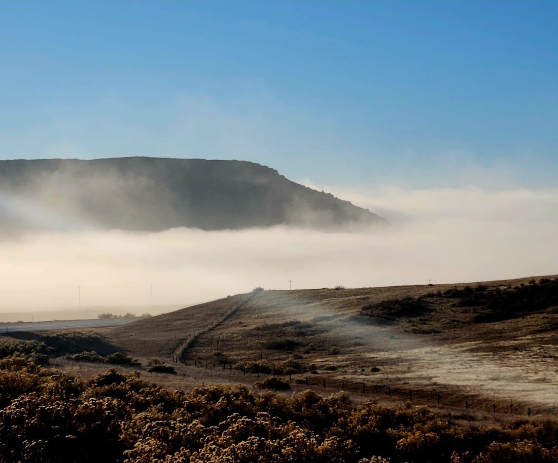 Low morning fog rolls below a flat-topped mesa above an open prairie and dirt road
