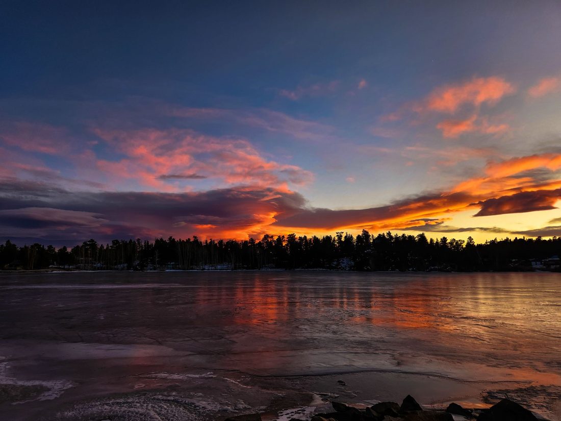 Pink and orange sunset clouds reflect across a frozen lake lined by a dark tree row