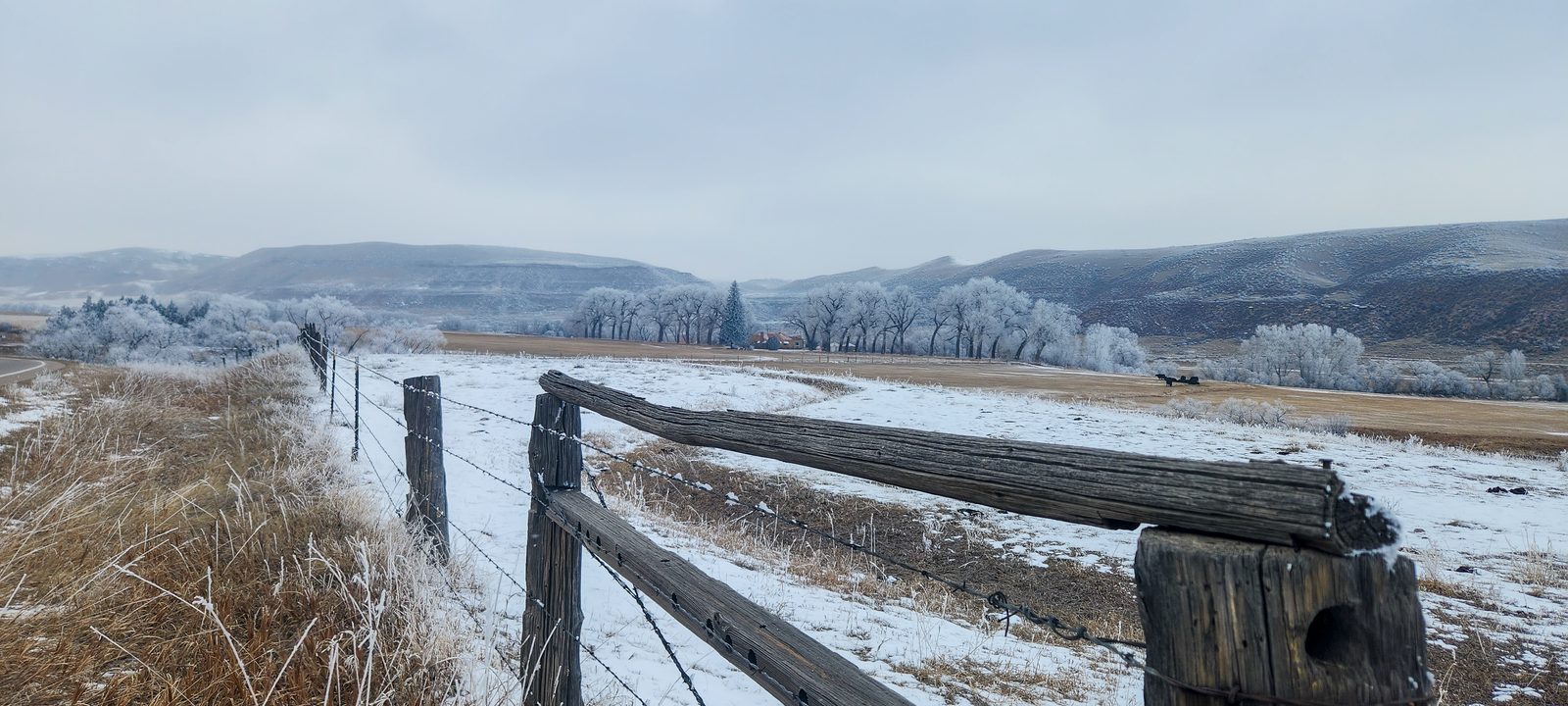 A weathered wooden ranch fence along a frosty snow-dusted pasture below low hills in small-town ranch country