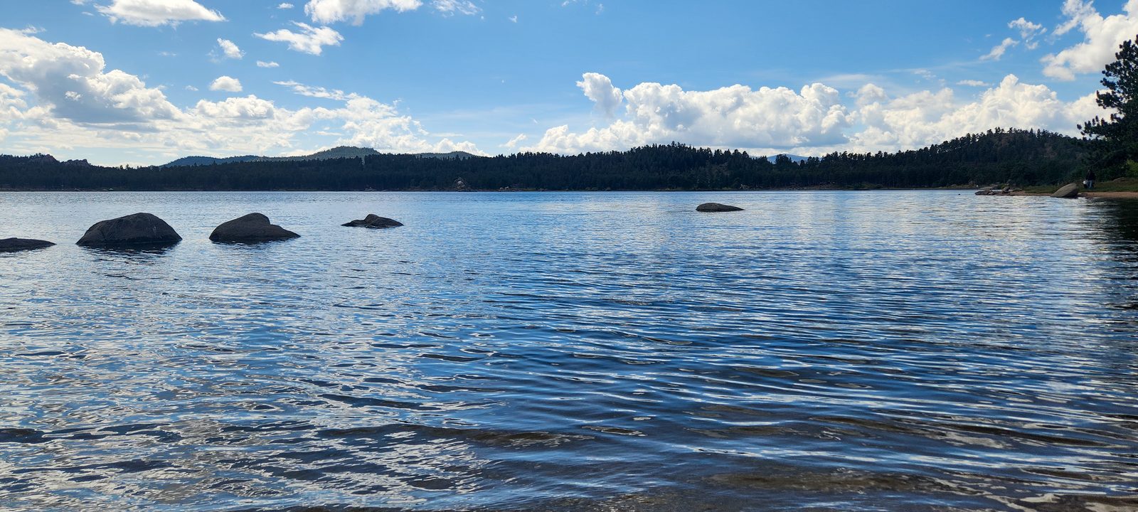 A sparkling blue mountain lake dotted with boulders below a forested ridge and summer clouds