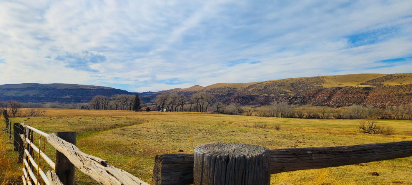 A weathered wooden fence along a golden open pasture with a lone ranch house tucked below the foothills