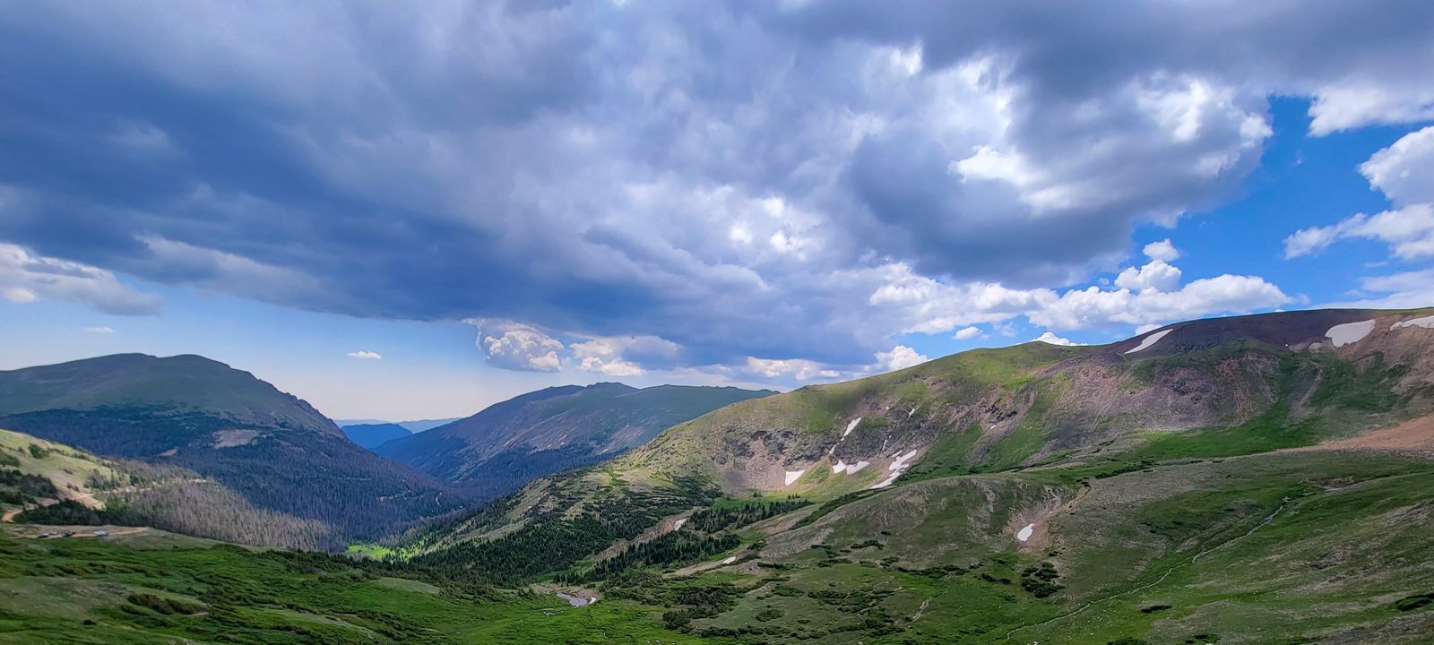 A sweeping high-country mountain vista of green alpine slopes and lingering snow under dramatic clouds